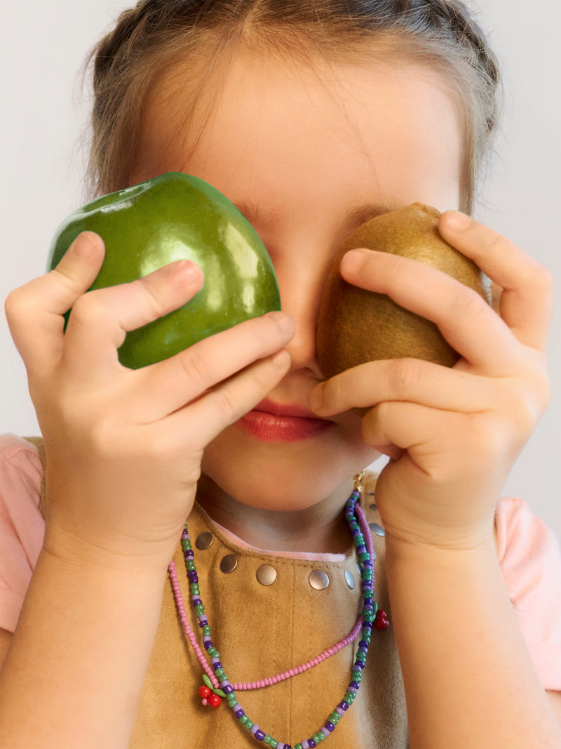 Young child holding a green bell pepper and brown kiwi fruit up to their eyes, wearing colorful beaded necklaces