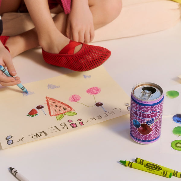 Child in red mesh shoes drawing with crayons on paper, with a purple drink can and colorful artwork visible on white surface.