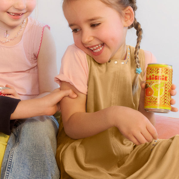 Two young girls smiling, one wearing beige overalls holding an orange and yellow can, the other in pink with pearl necklace