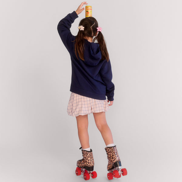 Young girl in navy hoodie and plaid skirt balancing golden cup on head while wearing leopard print roller skates with red wheels