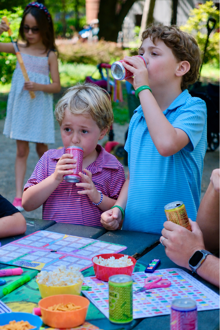 A group of children gathered around an outdoor picnic table playing a colorful board game with snacks and drinks. The scene is set in a sunny backyard or park with greenery in the background.
