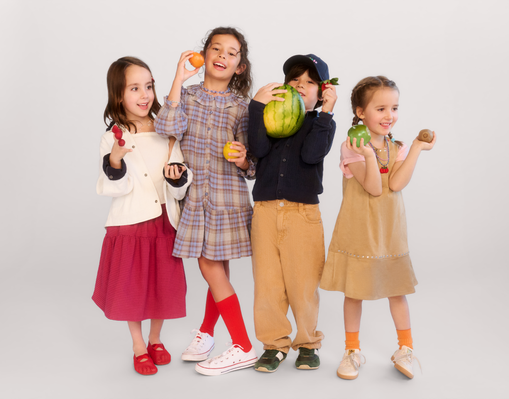Four children in colorful outfits holding various fruits including watermelon, orange, and apple against white background