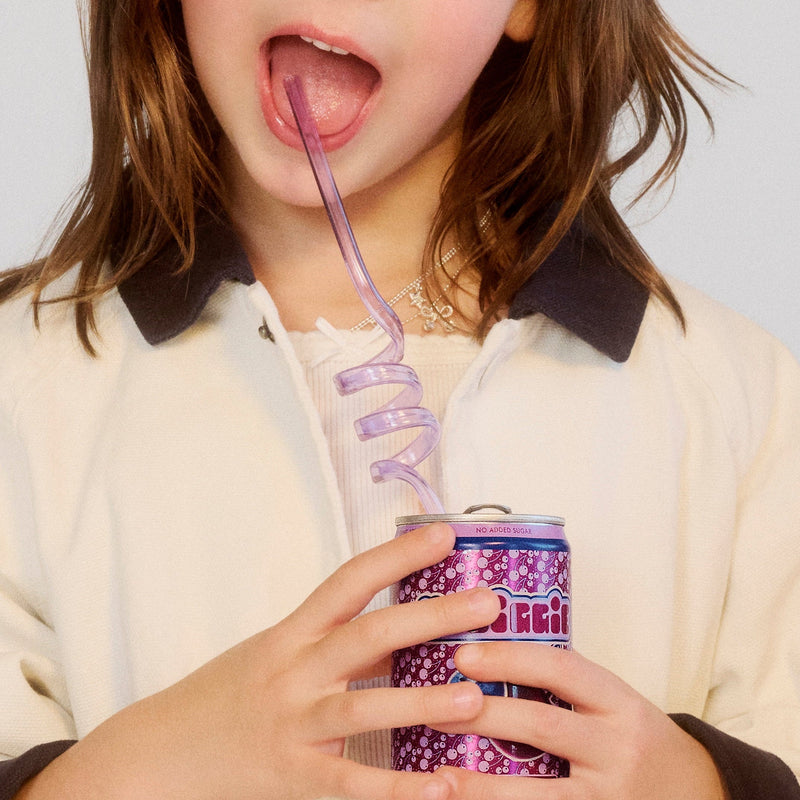 Woman with brown hair drinking from purple patterned can through curly purple straw, wearing cream hoodie with dark collar