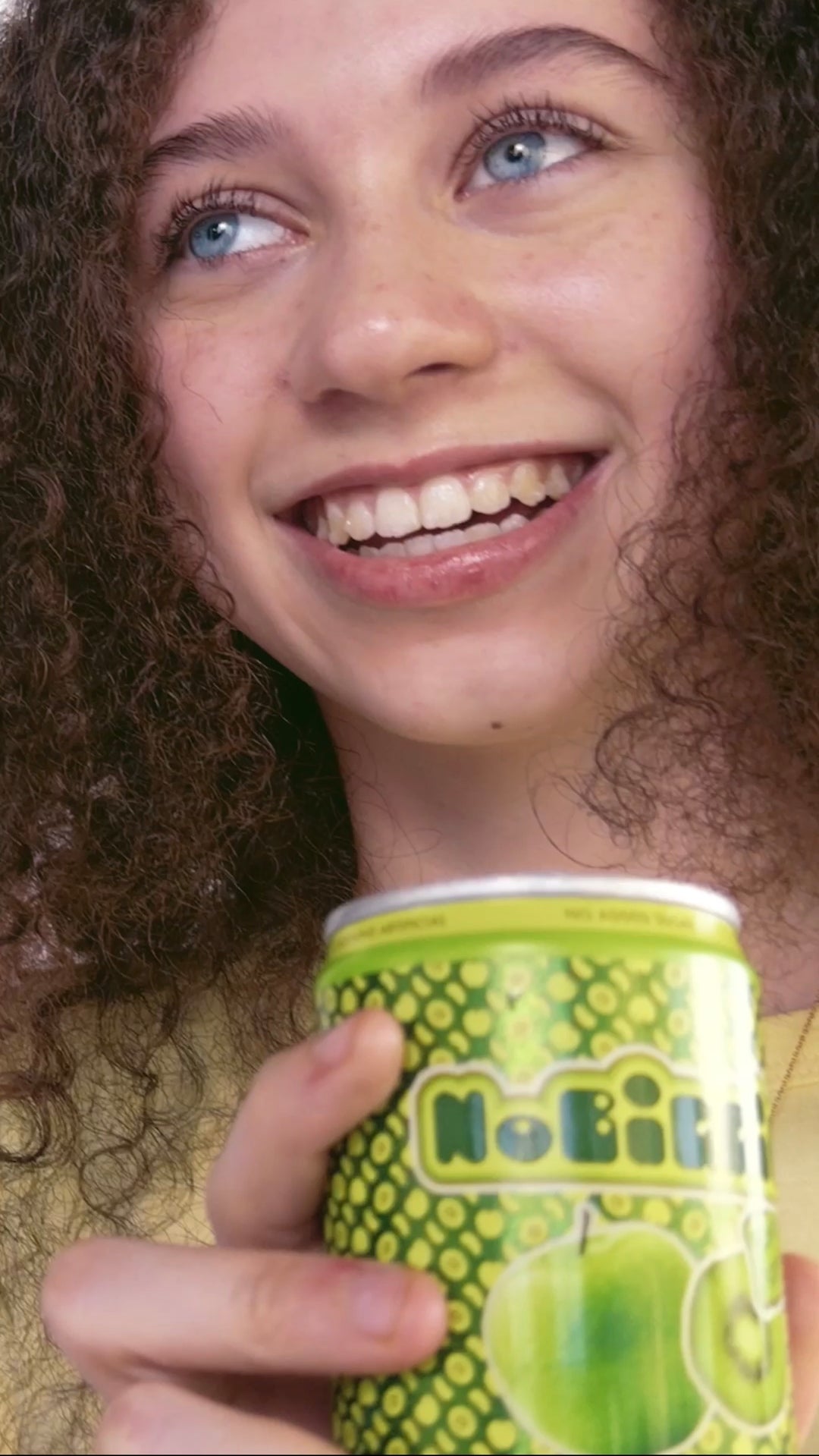 Smiling woman with curly hair and blue eyes holding a green "NoBiggie" branded can or cup with circular pattern design