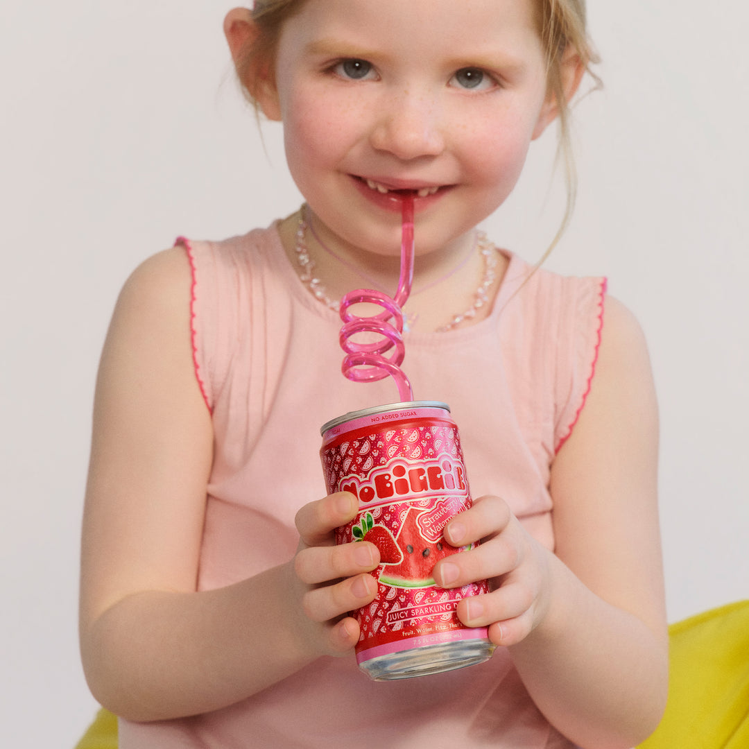 Young girl in pink dress drinking from red "Beehive" sparkling drink can with pink curly straw and pearl necklace