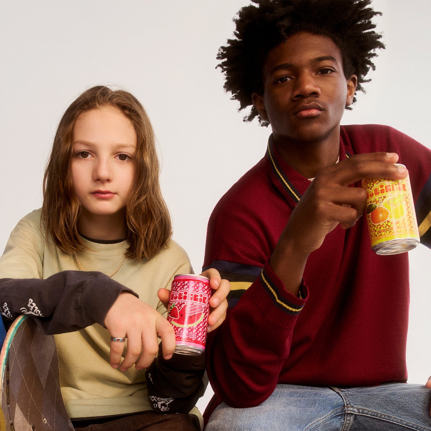 Two teenagers holding colorful drink cans - girl in beige hoodie with red can, boy in burgundy sweater with yellow can against white background