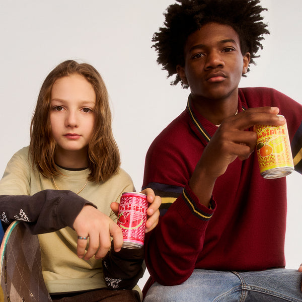 Two teenagers holding colorful drink cans - girl in beige hoodie with red can, boy in burgundy sweater with yellow can against white background