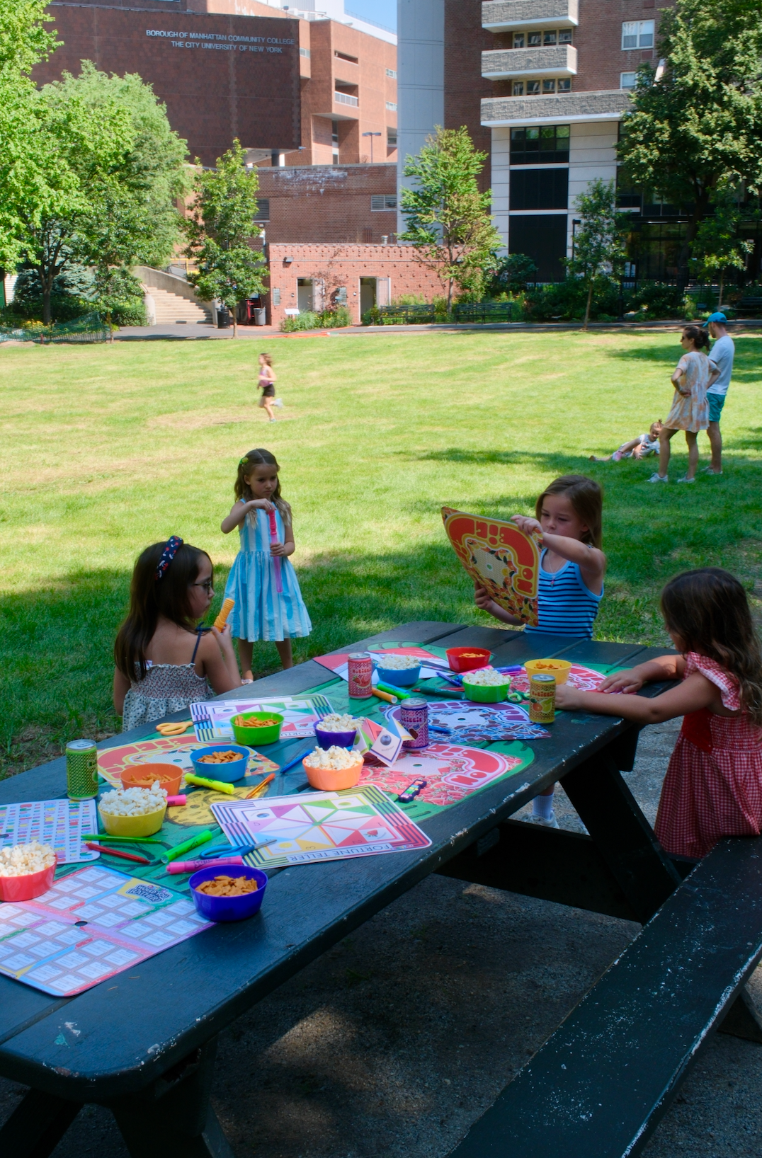 Children playing and socializing outdoors on a sunny day, with one girl holding a colorful book or card near a picnic table set with crayons, cups, and art supplies in a grassy park area with buildings in the background.