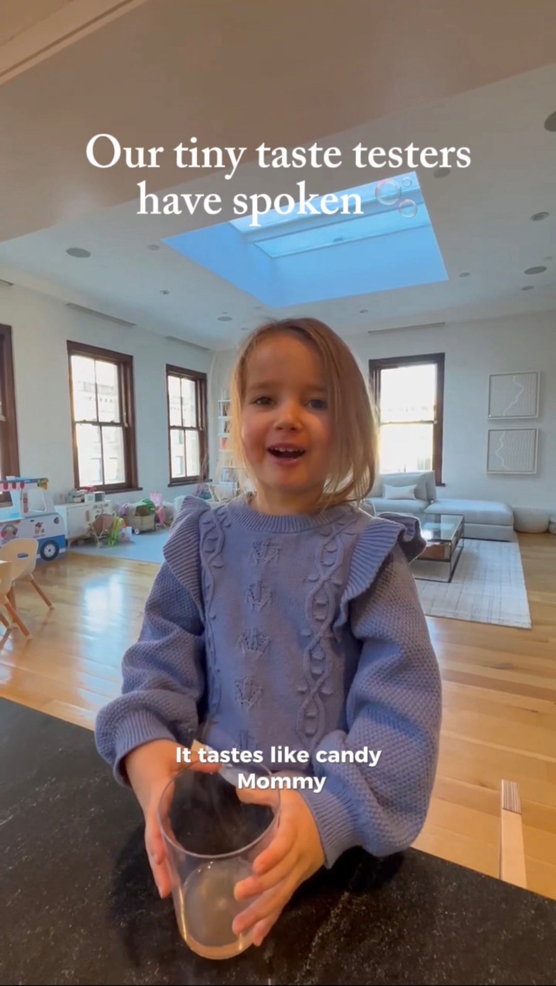 Young blonde girl in purple sweater holding glass with dark liquid in modern kitchen, text reads "Our tiny taste testers have spoken" and "It tastes like candy Mommy"